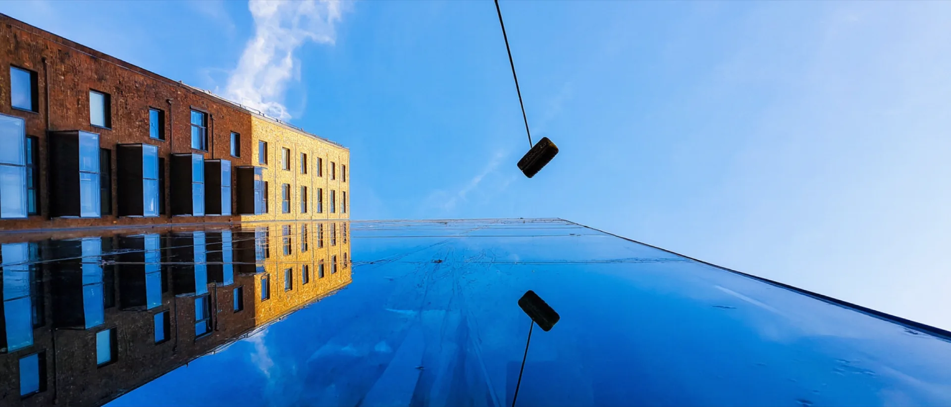 A brick and glass façade seen from below, pale blue sky mirrored in the panes, a window cleaner's pole crossing the frame